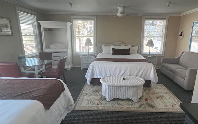A spacious hotel room with a queen bed center, white bedding, brown throw, and a wicker coffee table on a patterned rug.