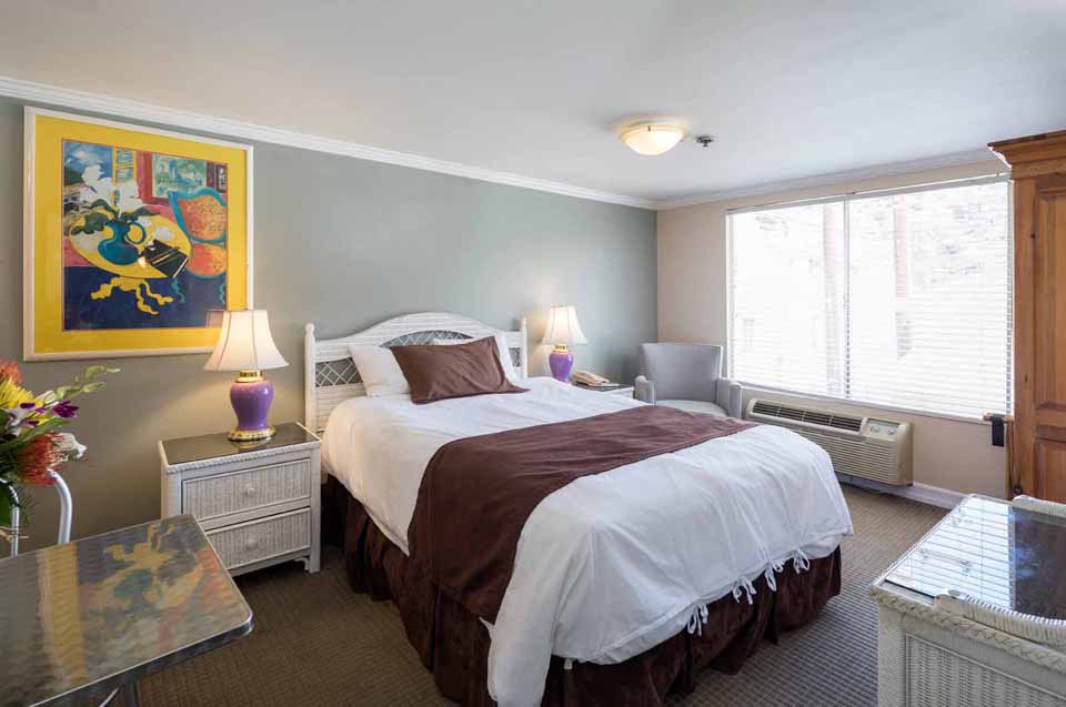 Hotel room with a white bed, brown accents, two matching nightstands and lamps, a colorful painting on the wall, and a large window with an air conditioner.